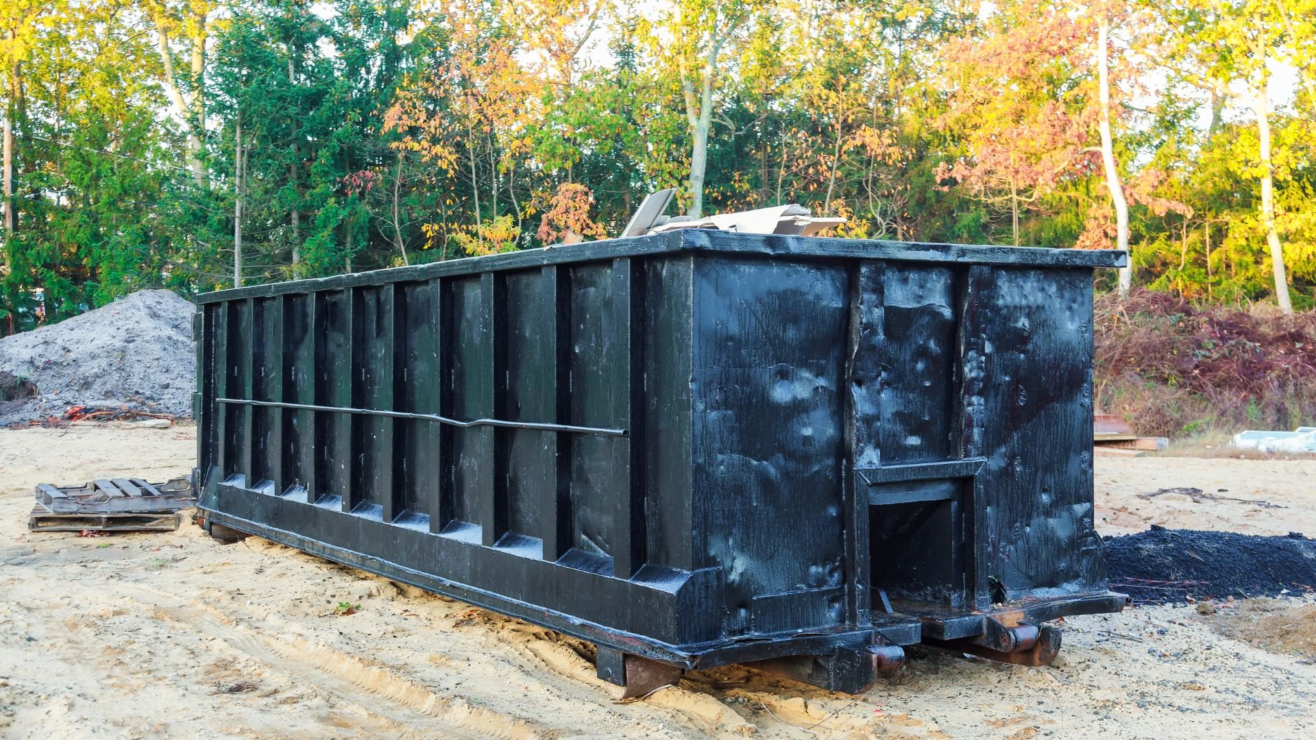 Large black dumpster on sandy ground with autumn trees in background