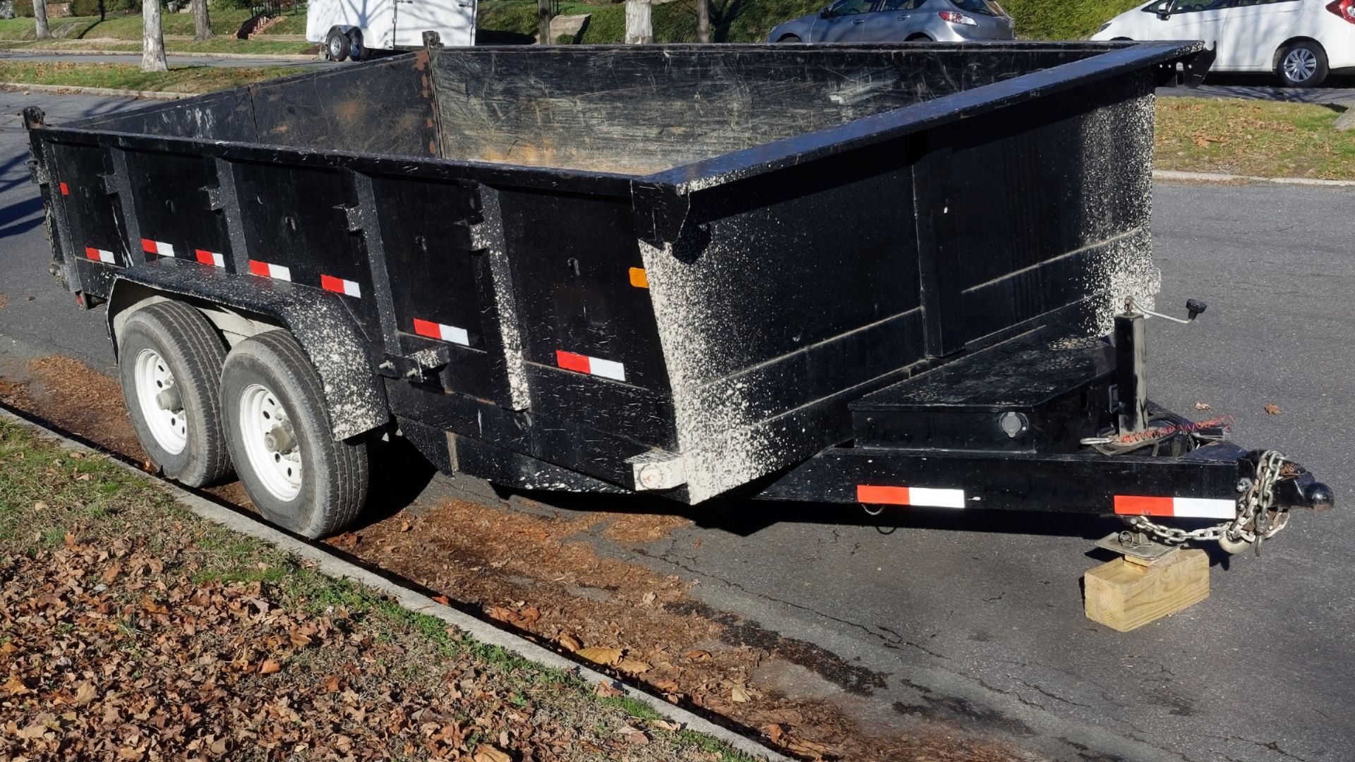 Black two-axle dump trailer parked on street with wooden block support