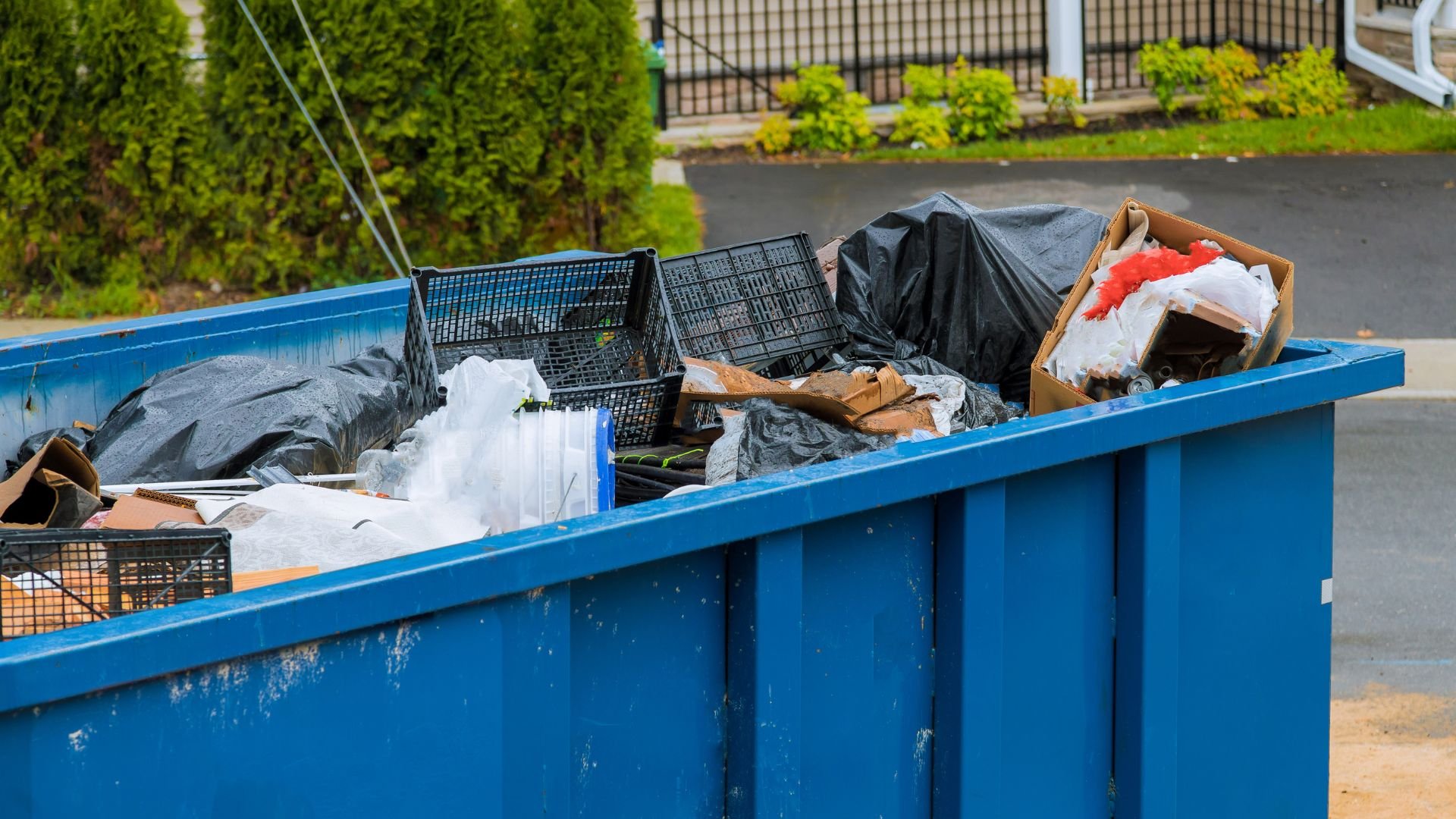 Blue dumpster filled with trash, plastic crates, and black garbage bags