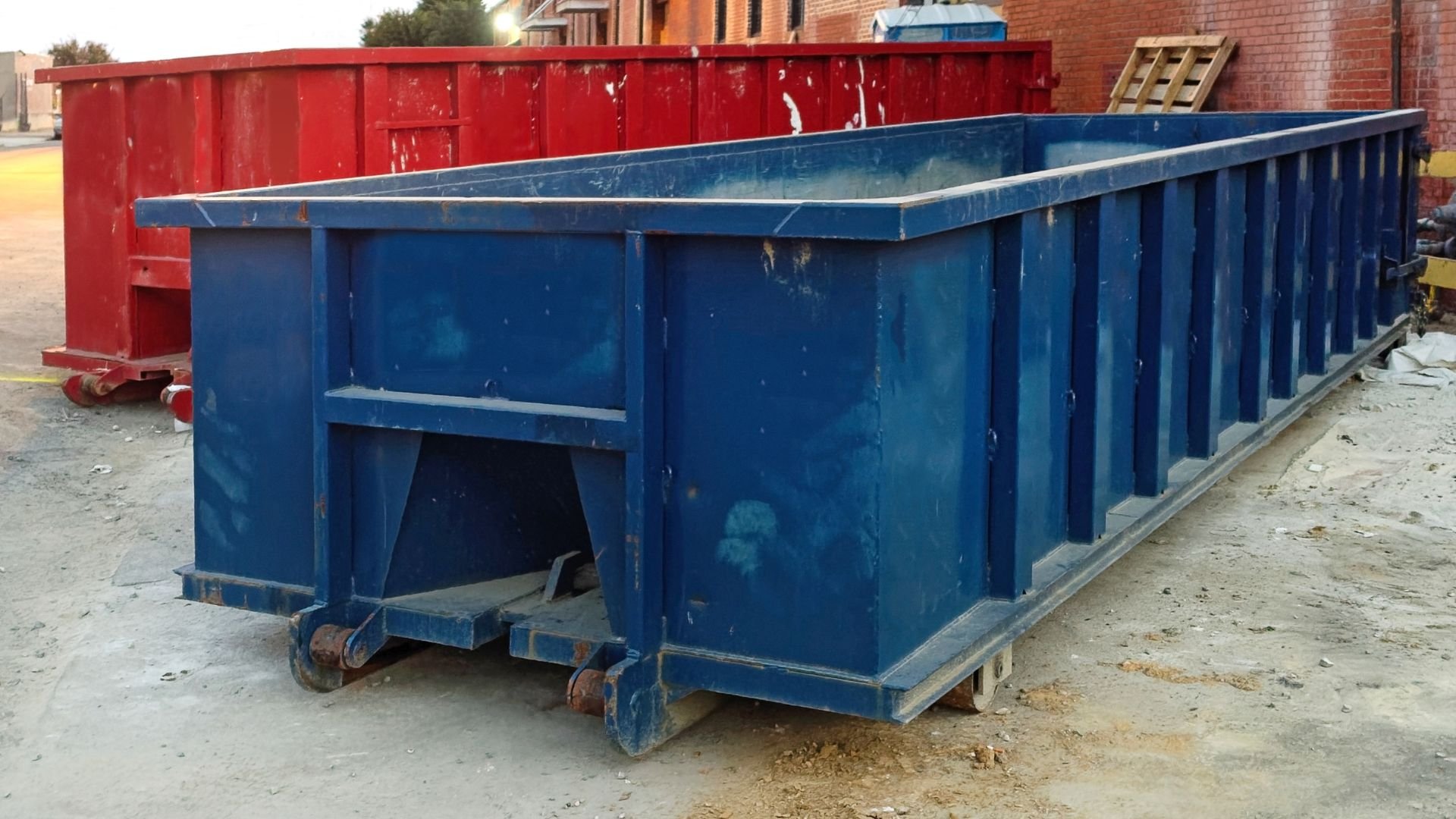 Blue and red industrial dumpsters on construction site with brick building